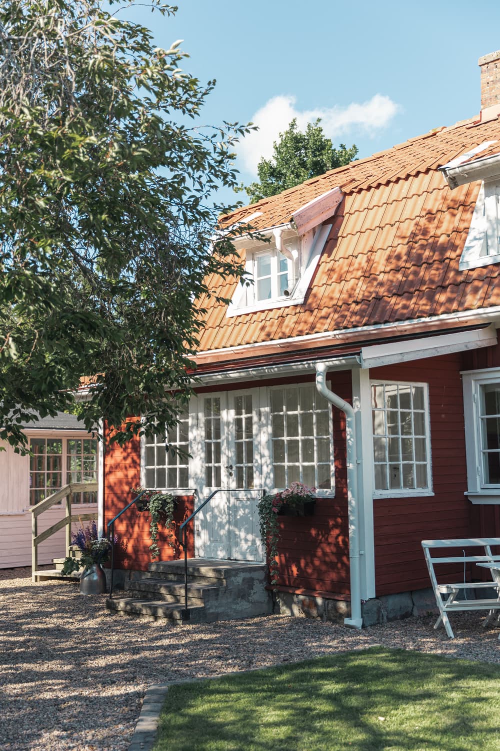 Gästhusets röda fasad och tegeltak under ett stort träd, med trädgårdsmöbler framför.
