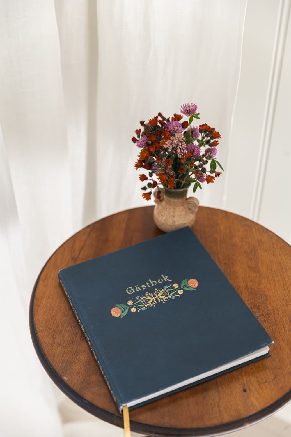 En blå gästbok och en bukett ängsblommor på ett runt träbord.