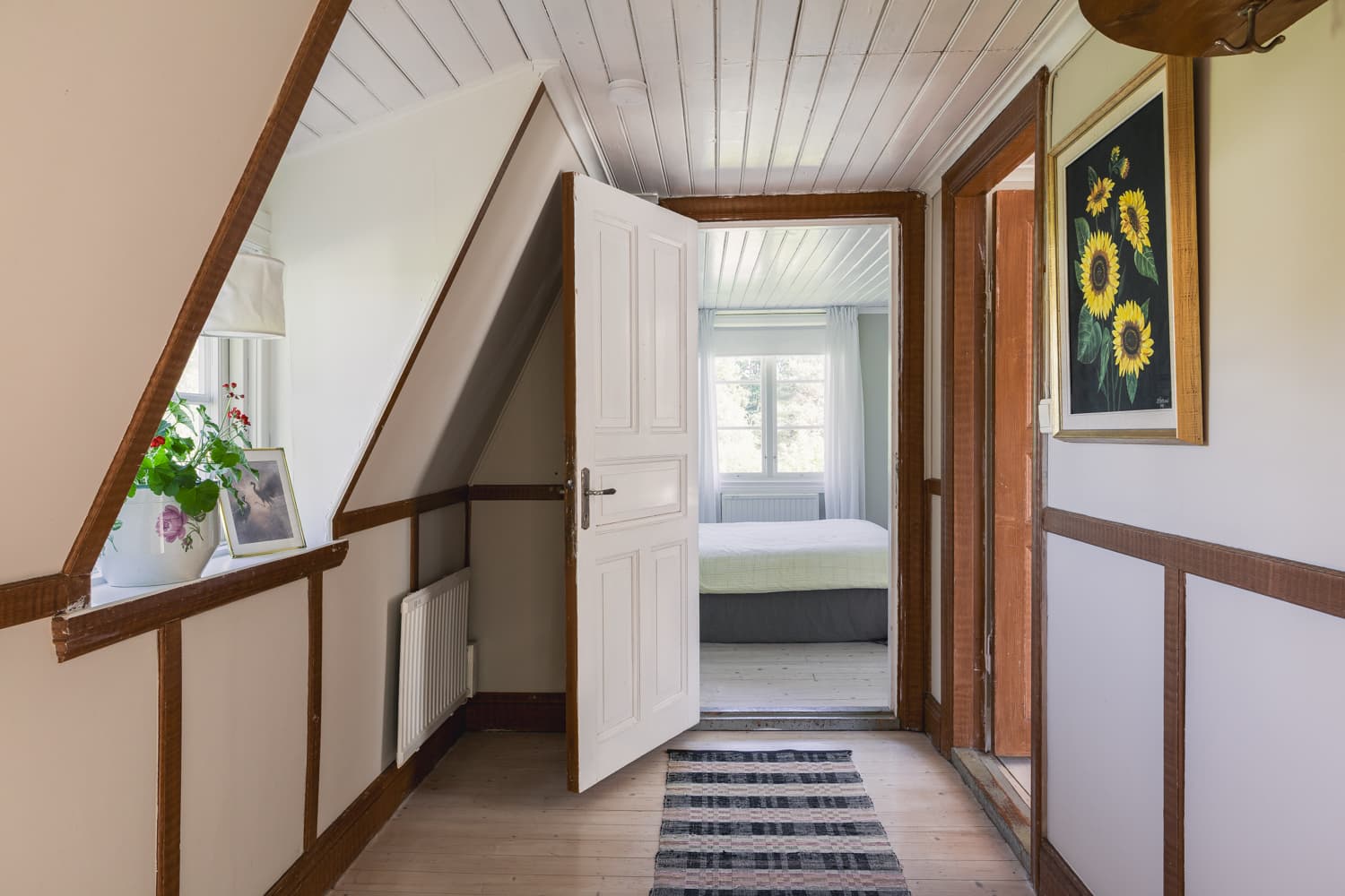 Gästhusets ljusa hall på övervåningen med snedtak, brunbetsade lister och en öppen dörr in mot ett sovrum.
