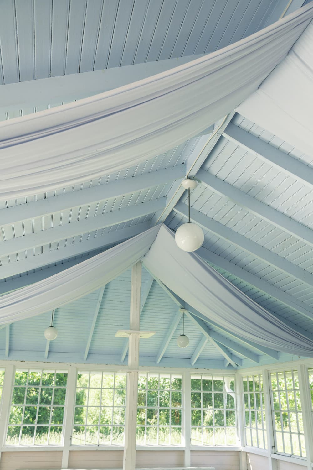 Ljusa tygvåder draperade under paviljongens blåmålade takbjälkar.