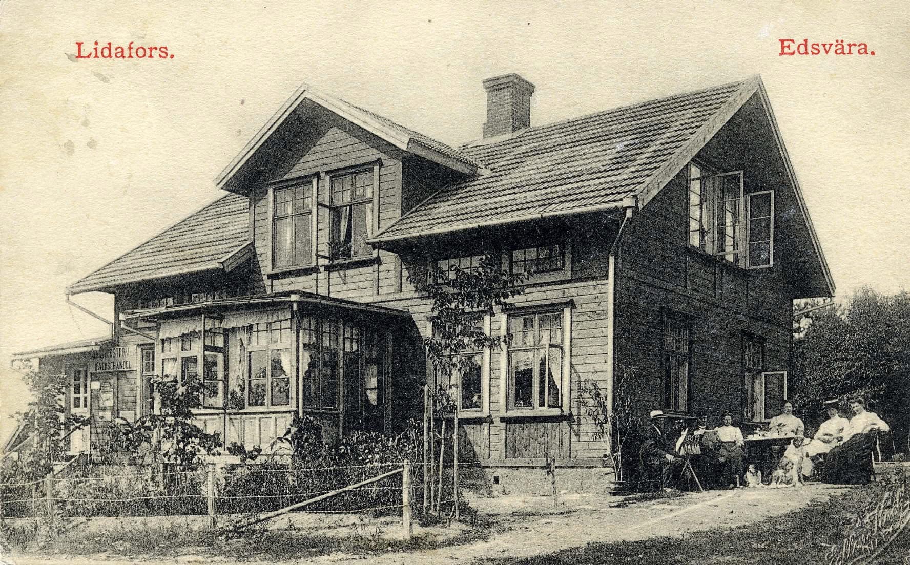 Gammalt svartvitt vykort märkt ”Lidafors. Edsvära.” som visar ett trähus med glasveranda, en skylt om diversehandel och ett sällskap vid ett bord i trädgården.