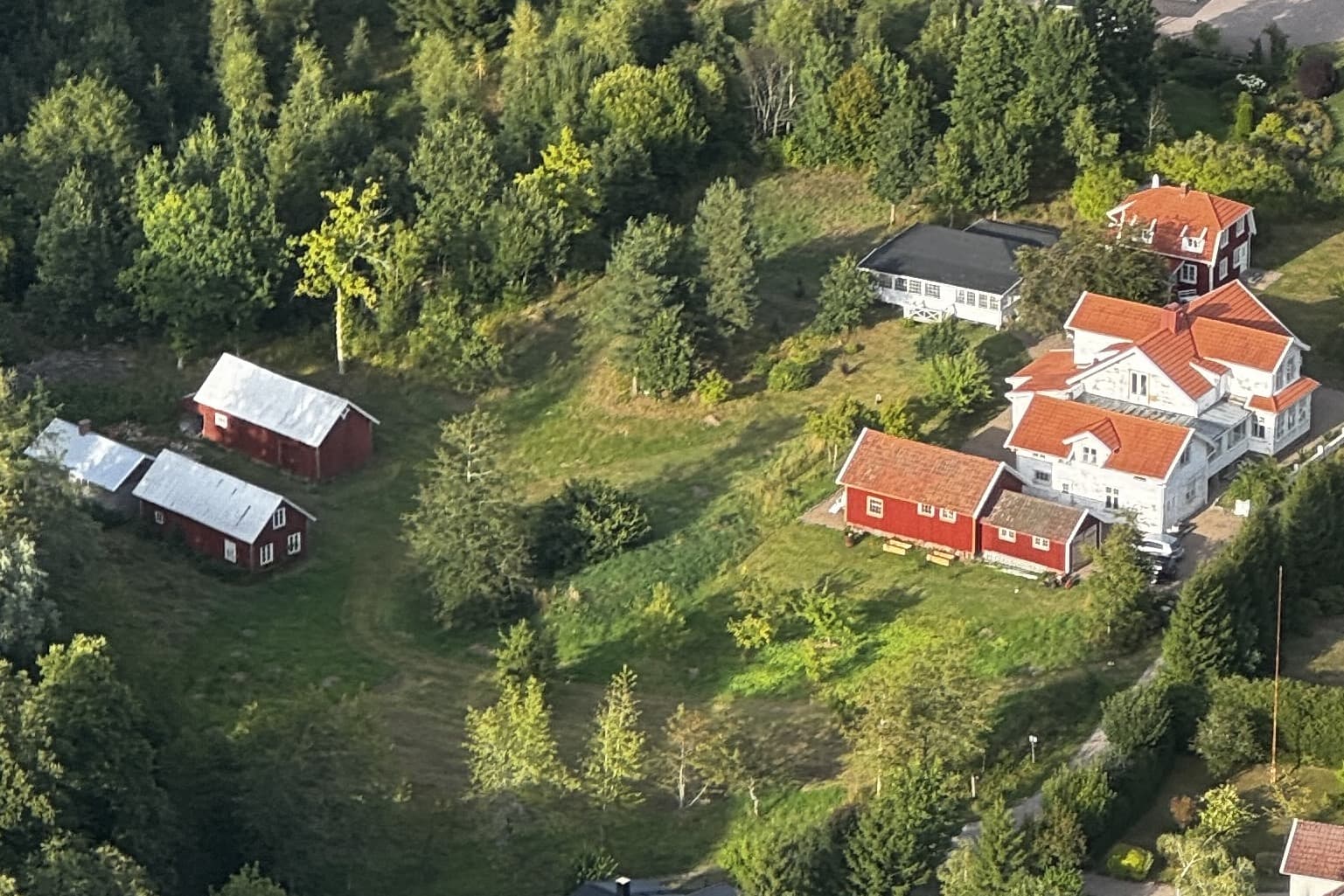 Flygbild över gården: det vita huset med orangea tegeltak, röda uthus med plåttak, en ljus byggnad med många fönster och en grön gårdsplan omgiven av träd.
