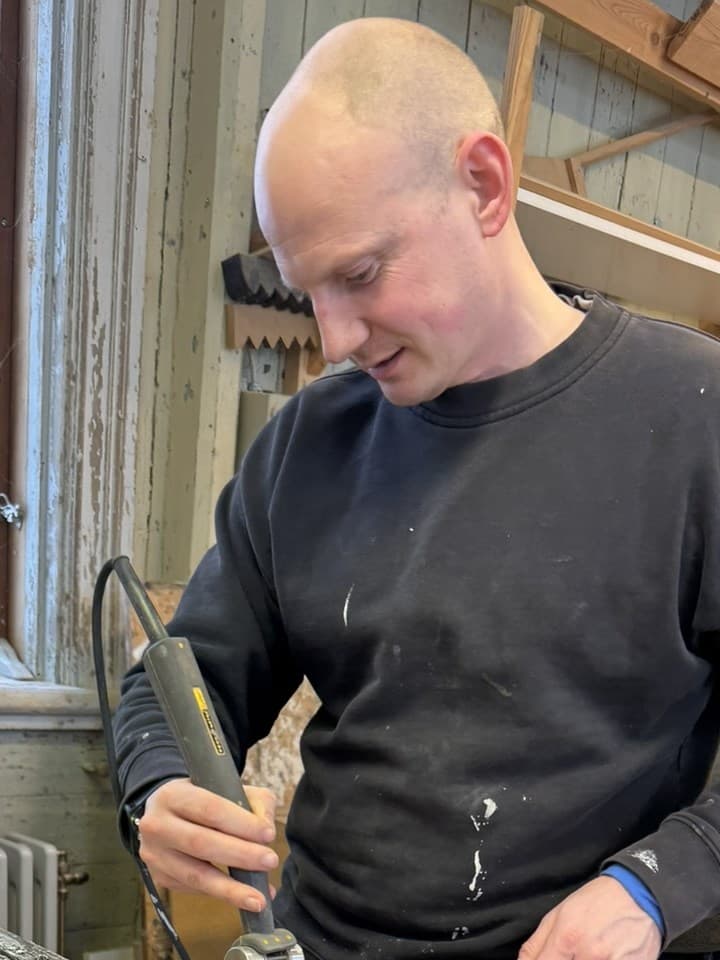 Lucas i mörk tröja skrapar bort gammal färg från en fönsterbåge med en värmeapparat och ett stämjärn.