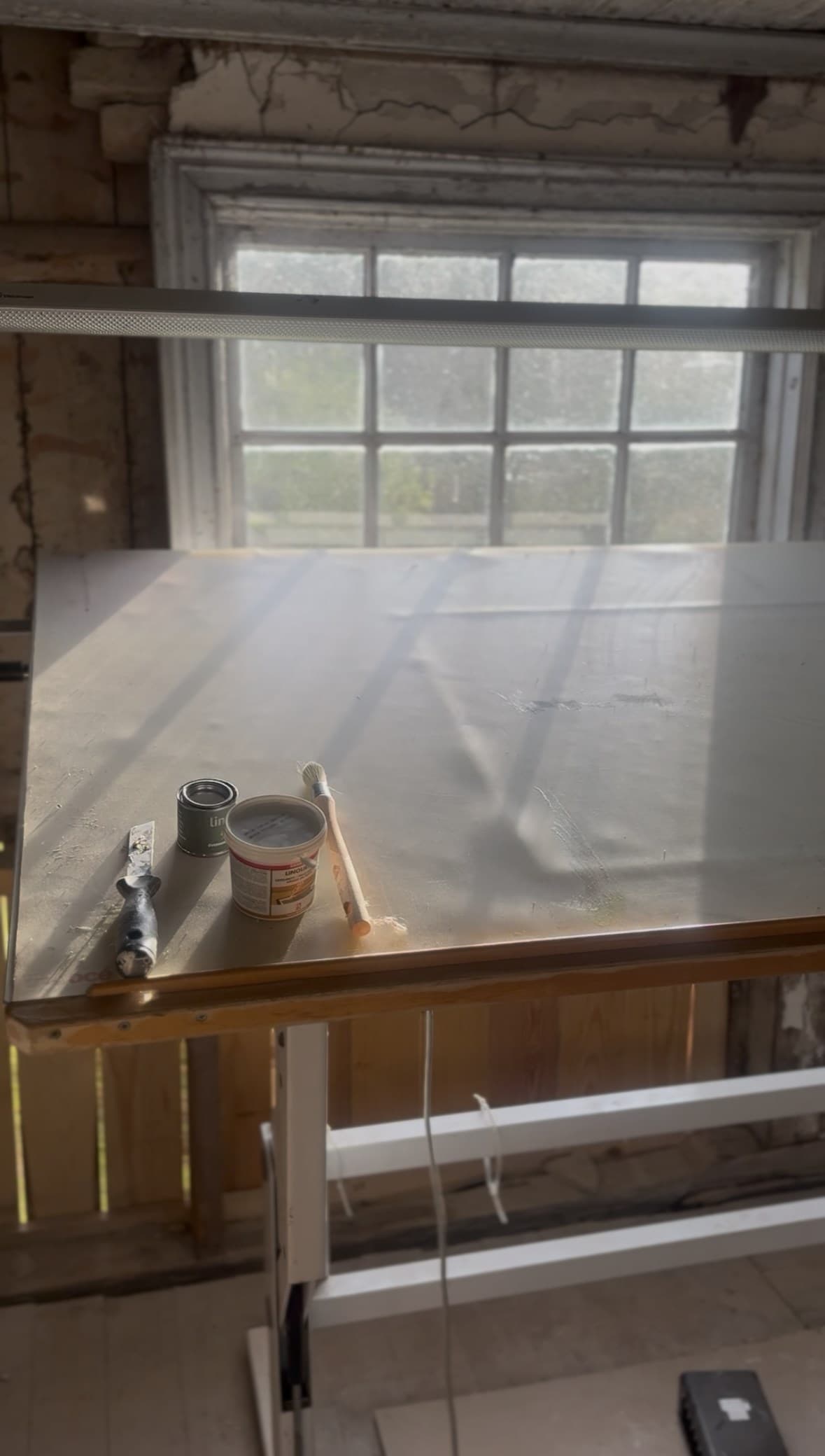 En kittkniv, burkar med linoljeprodukter och en pensel på en ljus arbetsbänk framför ett spröjsat fönster.