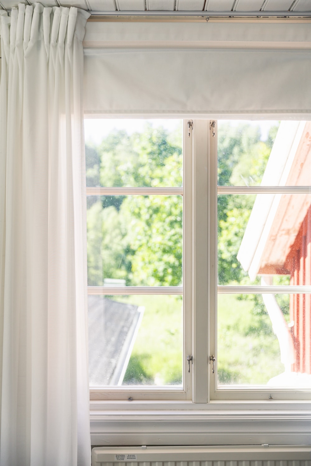 Utsikt genom ett vitt fönster mot träd och en röd husvägg.