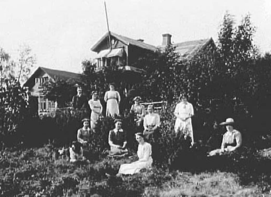 Ett tiotal kvinnor i ljusa klänningar och hattar sitter och står i en trädgård framför pensionatets byggnader.