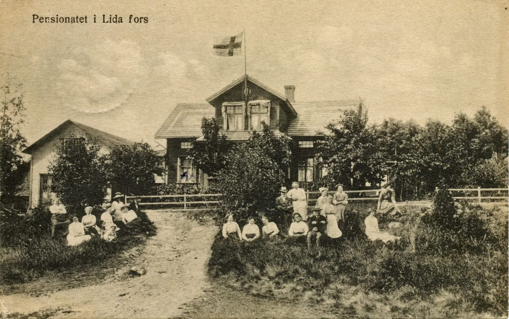 Vykort med texten ”Pensionatet i Lida fors”: ett trähus med flaggstång och sommargäster i ljusa kläder som sitter i gräset framför.