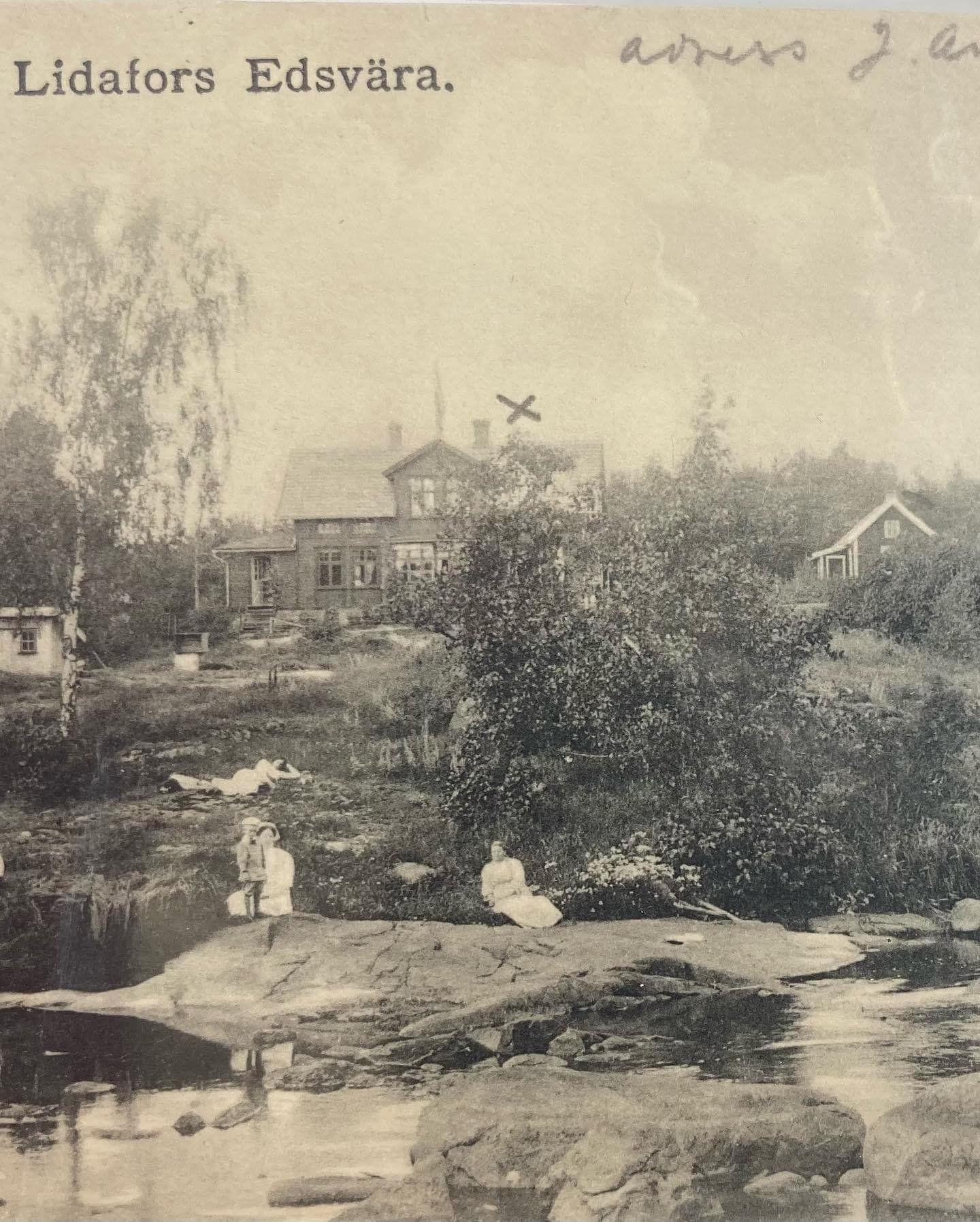 Vykort märkt ”Lidafors Edsvära.” med klipphällar och vatten i förgrunden, några personer i ljusa kläder vid ån och pensionatet bland träden bakom.